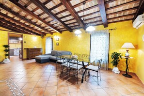 A room with a tiled floor, a sofa, a dining table with chairs, a lamp, and a ceiling with exposed beams.