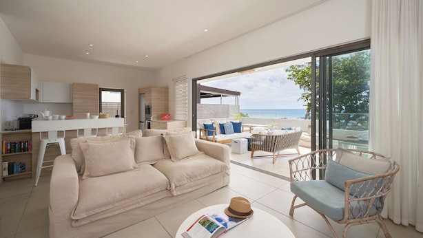 A modern living room with a sofa, a wicker chair, a dining table, and a view of the sea. - Les Estivales Beachfront Suites & Penthouses, LOV (NORTH)