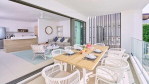 A modern dining area with a wooden table, white wicker chairs, and a view of the kitchen and living area. - Les Estivales Beachfront Suites & Penthouses, LOV (NORTH)