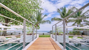 A wooden deck leading to a pool with white railings and lounge chairs, overlooking a beach and ocean. - Les Estivales Beachfront Suites & Penthouses, LOV (NORTH)