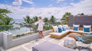 A balcony with a glass barrier, a sofa with cushions, a wicker chair, and a view of the ocean. - Les Estivales Beachfront Suites & Penthouses, LOV (NORTH)