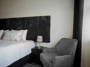 A hotel room with a bed, a nightstand, a lamp, a grey armchair, and a dark textured wall. - Room-king Deluxe Cottage Room (Cape Town)
