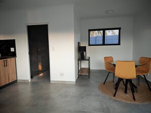 A modern kitchen with dark cabinetry, a dining area with two orange chairs and a glass table, and a window with blinds. - Room-king Deluxe Cottage Room (Cape Town)