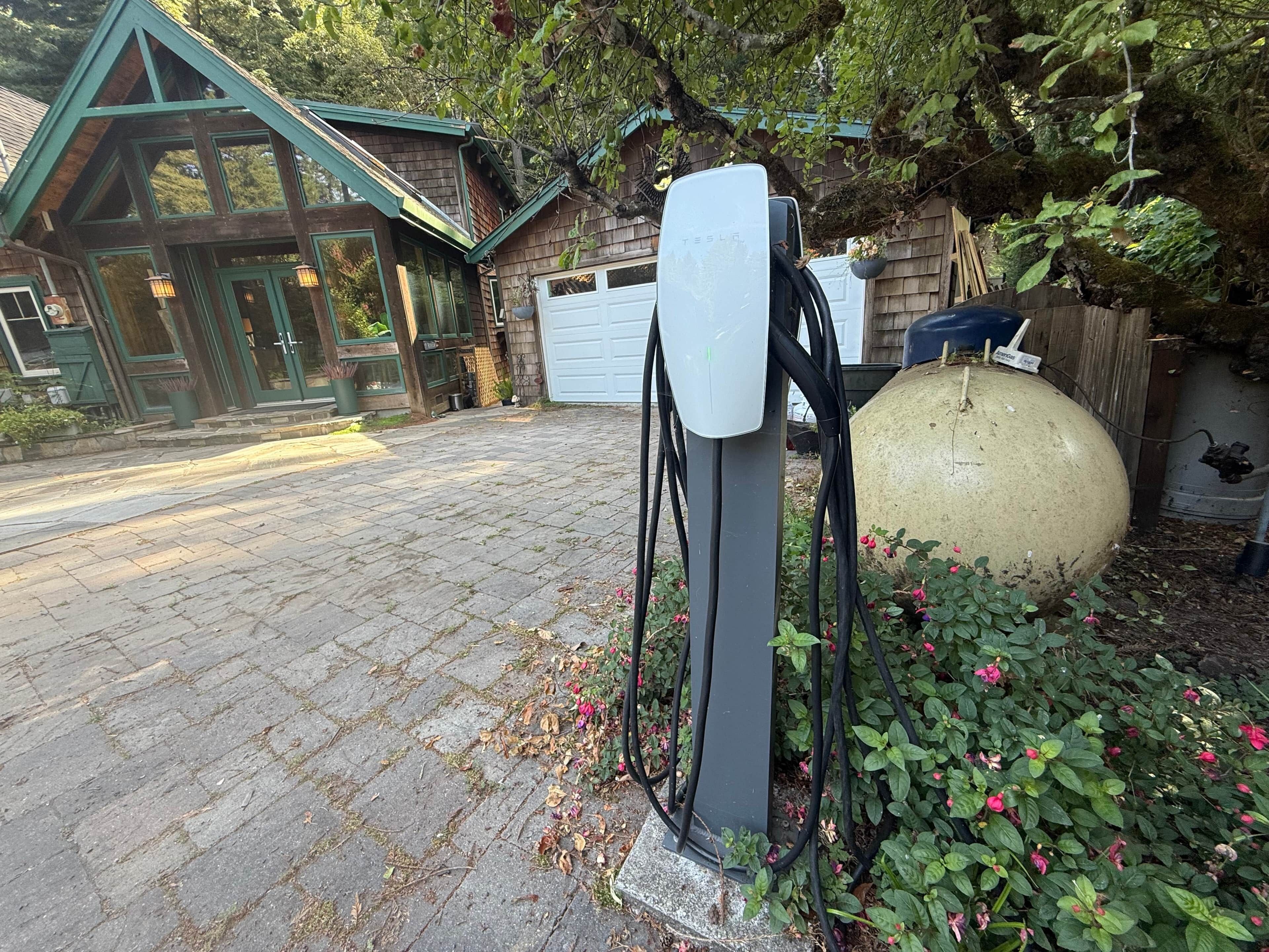 A Tesla charging station in a residential driveway with a brick house and a large decorative sphere in the background.
