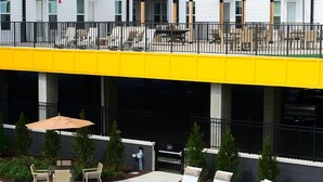 Un área de piscina con sillones de descanso y sombrillas, una terraza techada y un edificio con múltiples ventanas y balcones.