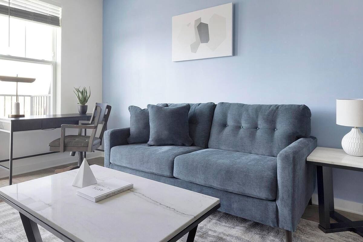 A living room with a grey sofa, a white coffee table, a side table with a lamp, a framed picture on the wall, and a window with blinds.