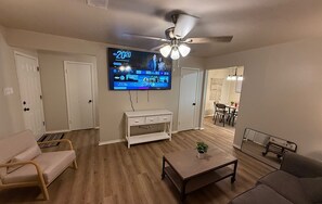 Ein Wohnzimmer mit Fernseher, Deckenventilator, weißen Möbeln und einem Essbereich im Hintergrund.