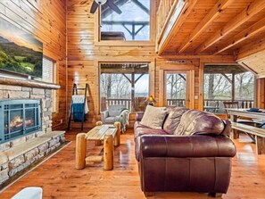 A log cabin interior with a stone fireplace, leather sofa, wooden dining table, and a view of the outdoors. - Sevierville Cabin w/ Views, Hot Tub, Pool Table & Resort Pool (Sevierville)