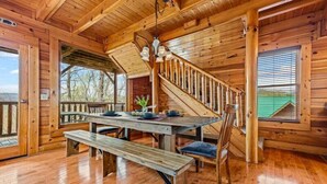 Ein Holzblockhaus-Interieur mit Esstisch, Bänken, einer Treppe und Blick durch ein Fenster.