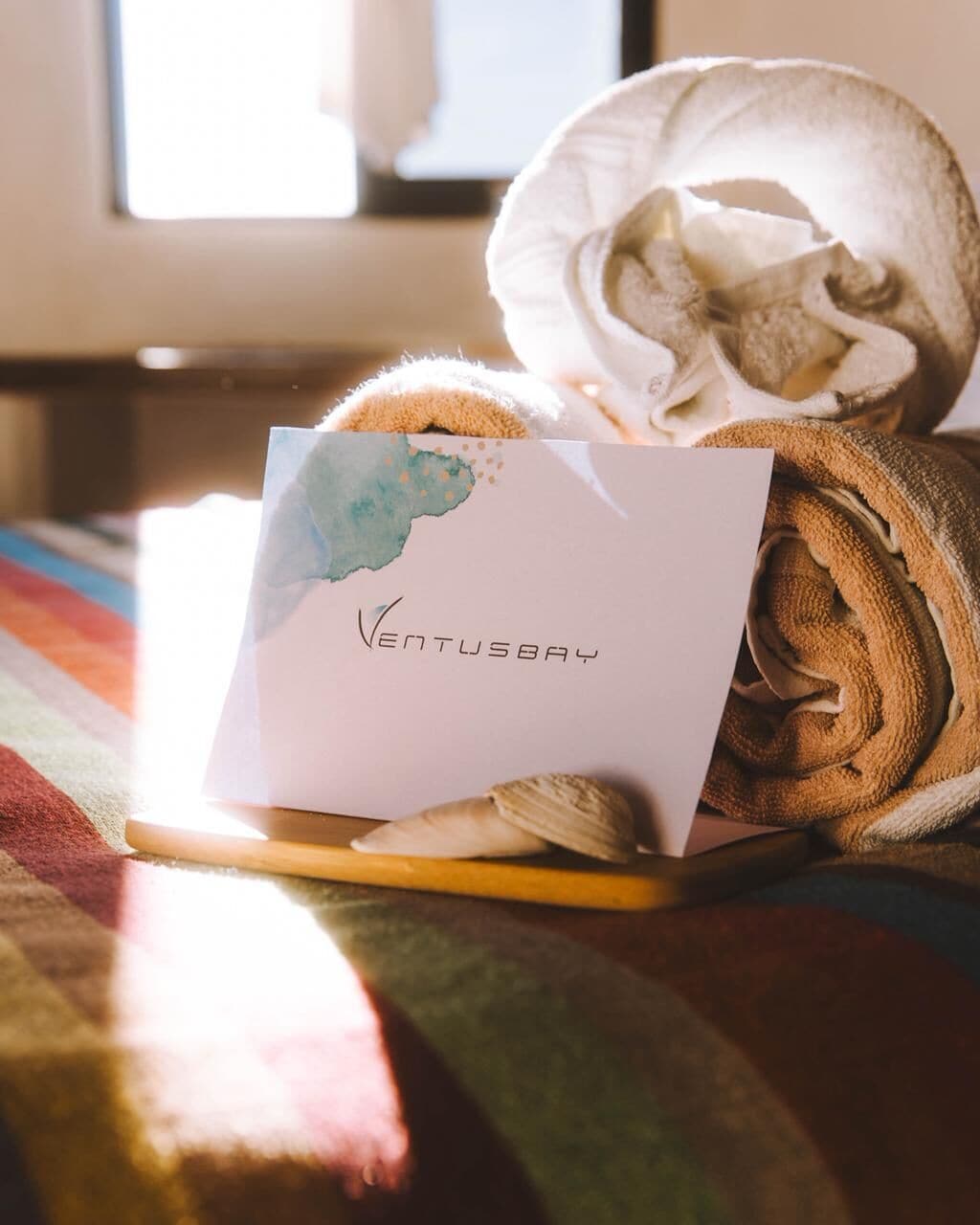 A neatly rolled towel, a card with 'VENTUSBAY' written on it, and a wooden tray on a bed with a colorful blanket.