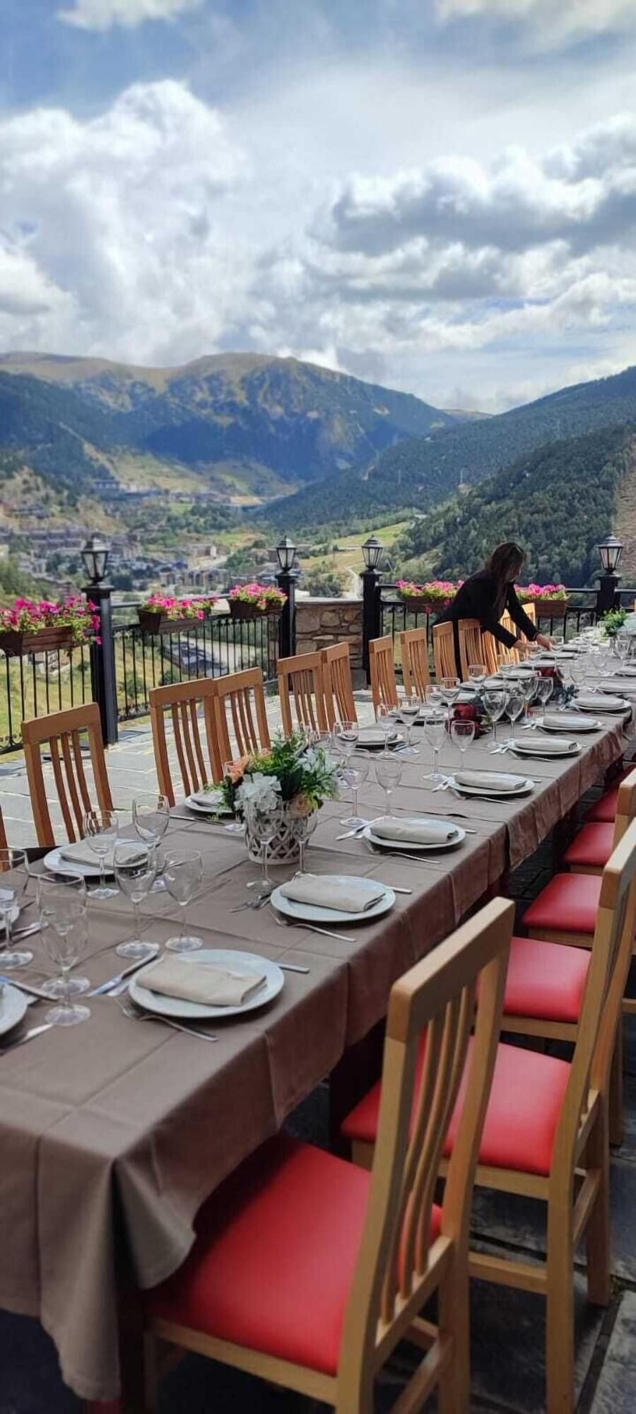 Una larga mesa de comedor al aire libre con sillas, vistas a montañas y cielo nublado.