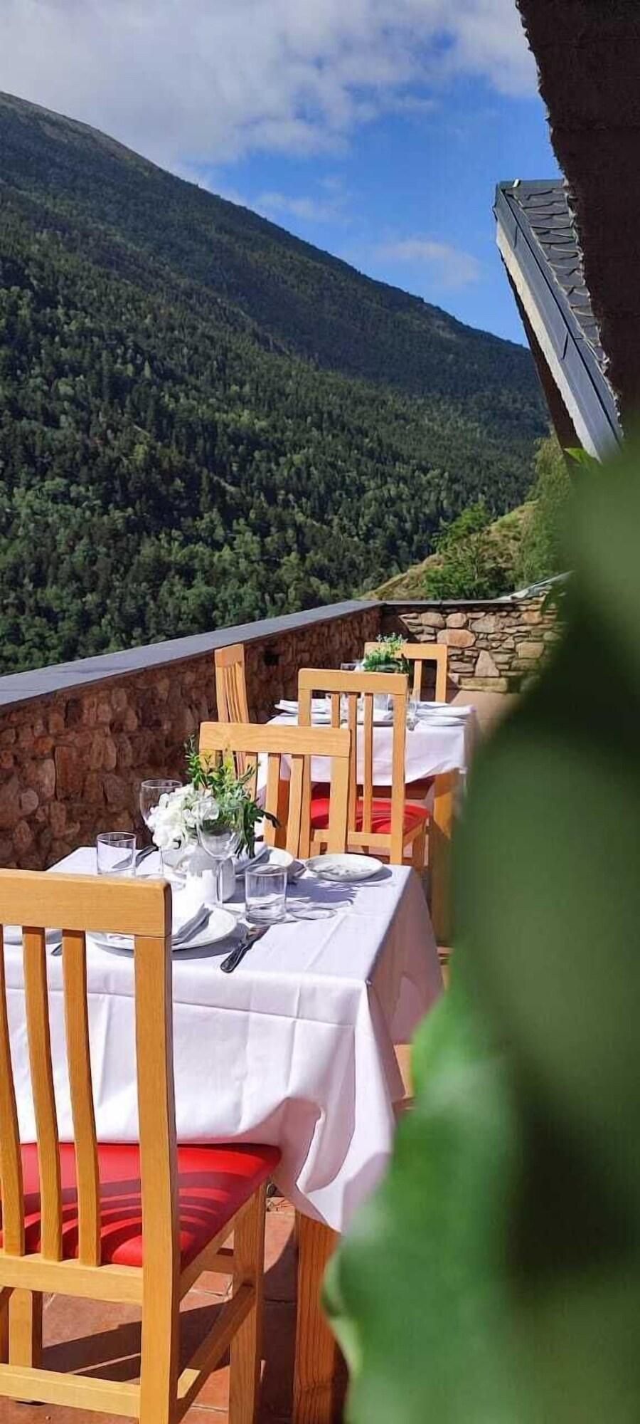 Un restaurante con terraza al aire libre con vistas a una montaña boscosa. Las mesas están dispuestas con manteles blancos y cojines rojos.