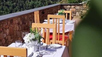 Un restaurante con terraza al aire libre con vistas a una montaña boscosa. Las mesas están dispuestas con manteles blancos y cojines rojos.