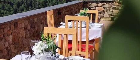 A restaurant with outdoor seating overlooking a forested mountain. Tables are set with white tablecloths and red cushions.