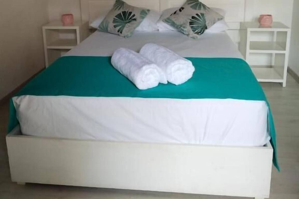 A neatly made bed with white bedding and a teal accent, two pillows with leaf patterns, and a small bedside table with a pink candle.