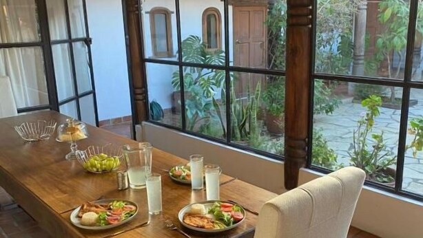 A dining area with a long wooden table, multiple chairs, hanging pendant lights, and a view of a garden through large windows. - Arles ByB Cafe (San Cristóbal de las Casas)