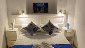 A neatly made bed with a blue bedspread and two pillows, a white headboard, and a nightstand with a lamp. - Arles ByB Cafe (San Cristóbal de las Casas)
