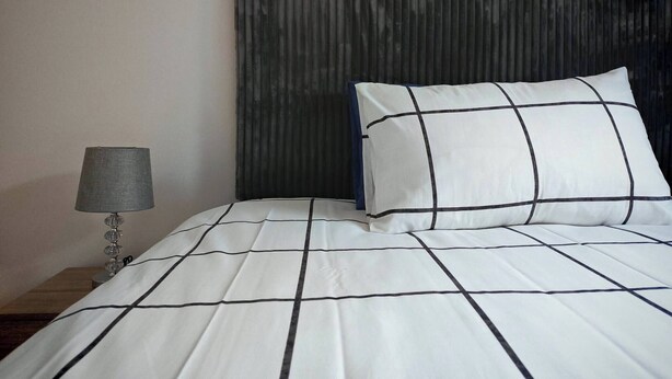 A bed with a white and black grid-patterned bedspread, a single pillow with a similar pattern, a dark headboard, and a bedside lamp with a grey shade. - Room-courtyard Room With Shared Bathroom (Cape Town)