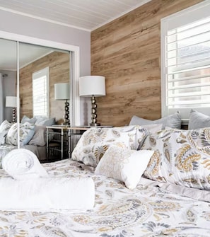 A bedroom with a large bed, a mirror, a bedside table with a lamp, and a window with blinds.