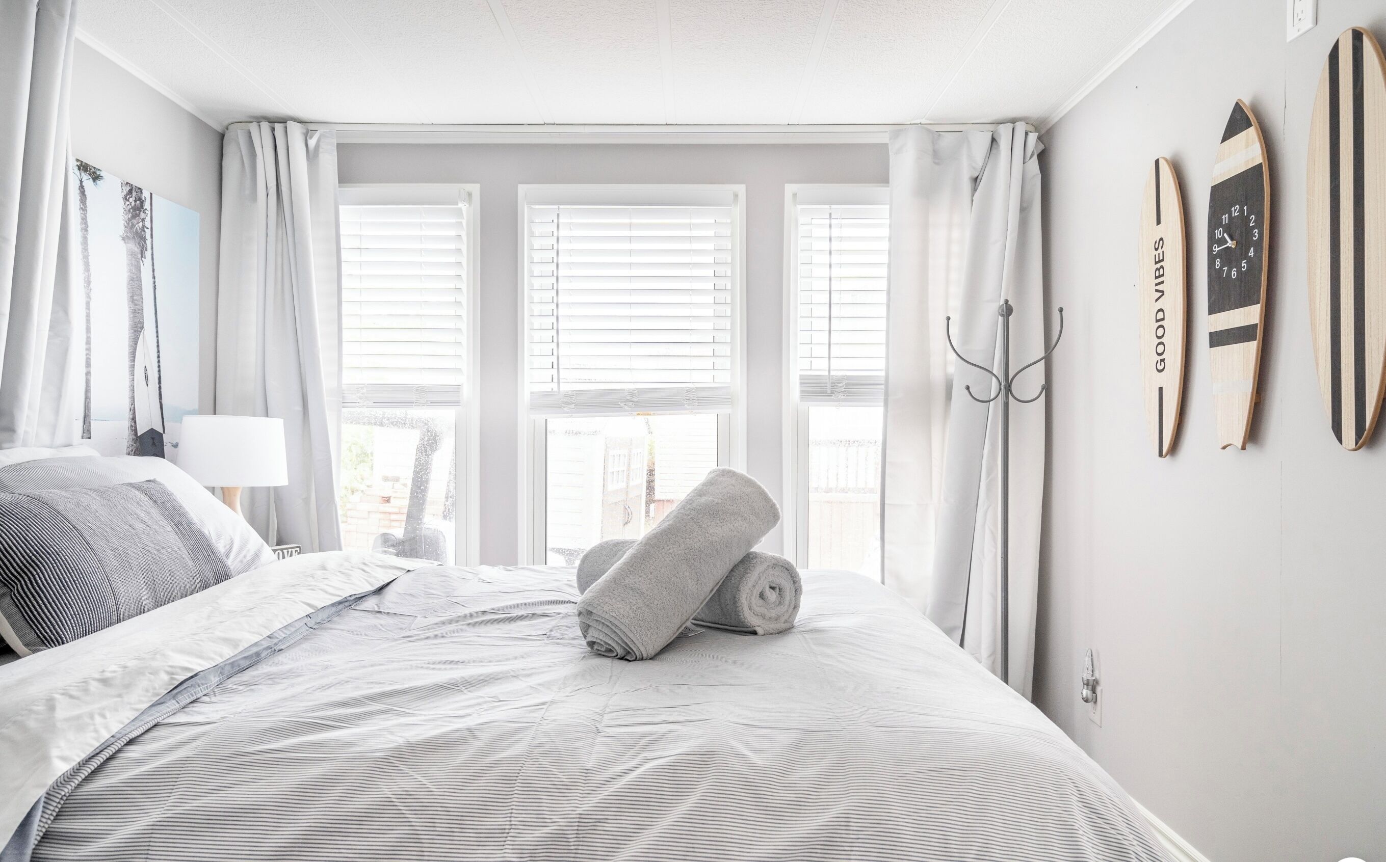 Una camera d'albergo con un letto, una finestra con persiane, un asciugamano sul letto e un appendiabiti con un surfboard e una sveglia.