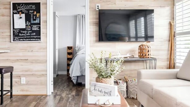 Un salon moderne avec un plancher en bois, un pouf en cuir marron, un canapé blanc, un téléviseur à écran plat fixé au mur et une pancarte « Bienvenue » accrochée au mur.