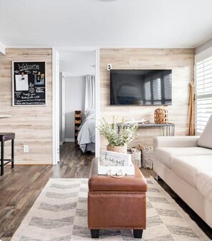 A modern living room with a wooden floor, a brown leather ottoman, a white sofa, a flat-screen TV mounted on the wall, and a 'Welcome' sign on the wall.