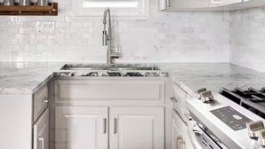 Une cuisine avec des meubles blancs, une fenêtre avec des stores, un évier en acier inoxydable et une cuisinière à gaz.