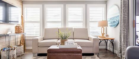Una sala de estar con un sofá color beige, un reposapiés marrón, una mesita auxiliar con una lámpara y un televisor montado en la pared.
