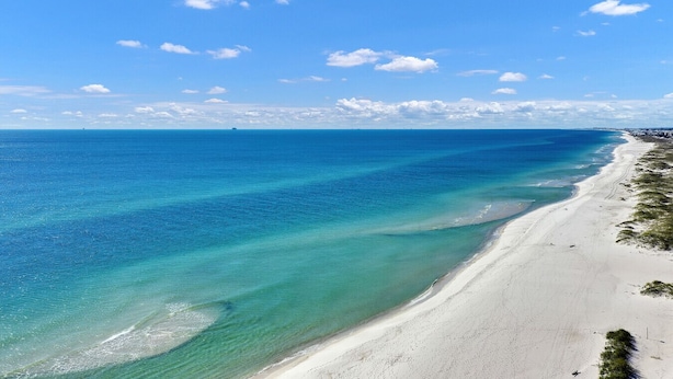 Une plage à l’eau turquoise et limpide, un rivage sablonneux et, au loin, la silhouette d’une ville.