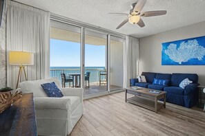 A room with a white sofa, a blue sofa, a coffee table, a ceiling fan, and a large window with a view of the ocean. - The Beach Club 1 Bedroom (Gulf Shores)