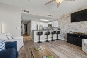 A modern living room with a sofa, coffee table, and a kitchen area with bar stools. - The Beach Club 1 Bedroom (Gulf Shores)