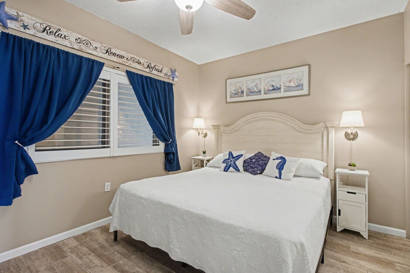 A bedroom with a bed, two bedside tables, a window with curtains, a ceiling fan, and a wall-mounted artwork.