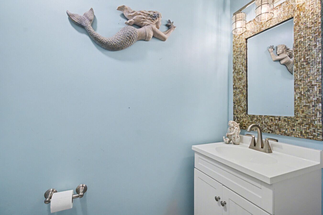 A bathroom with a light blue wall, a white vanity, a mirror with mosaic tile, and a mermaid decoration.