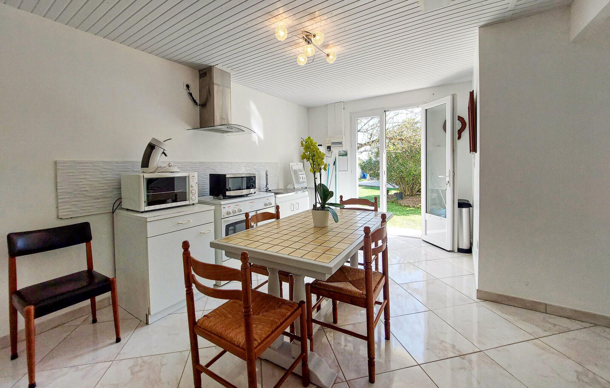 Een keuken met betegelde vloer, witte kasten, een eettafel met stoelen en uitzicht door een glazen deur.