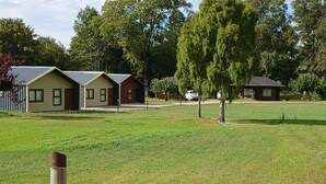 A row of single-story buildings with a flat roof, surrounded by a grassy field and trees.