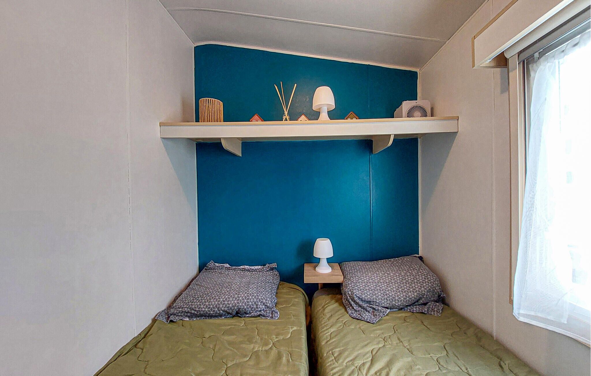 A room with two beds, a blue accent wall, a shelf with a lamp, and a window.