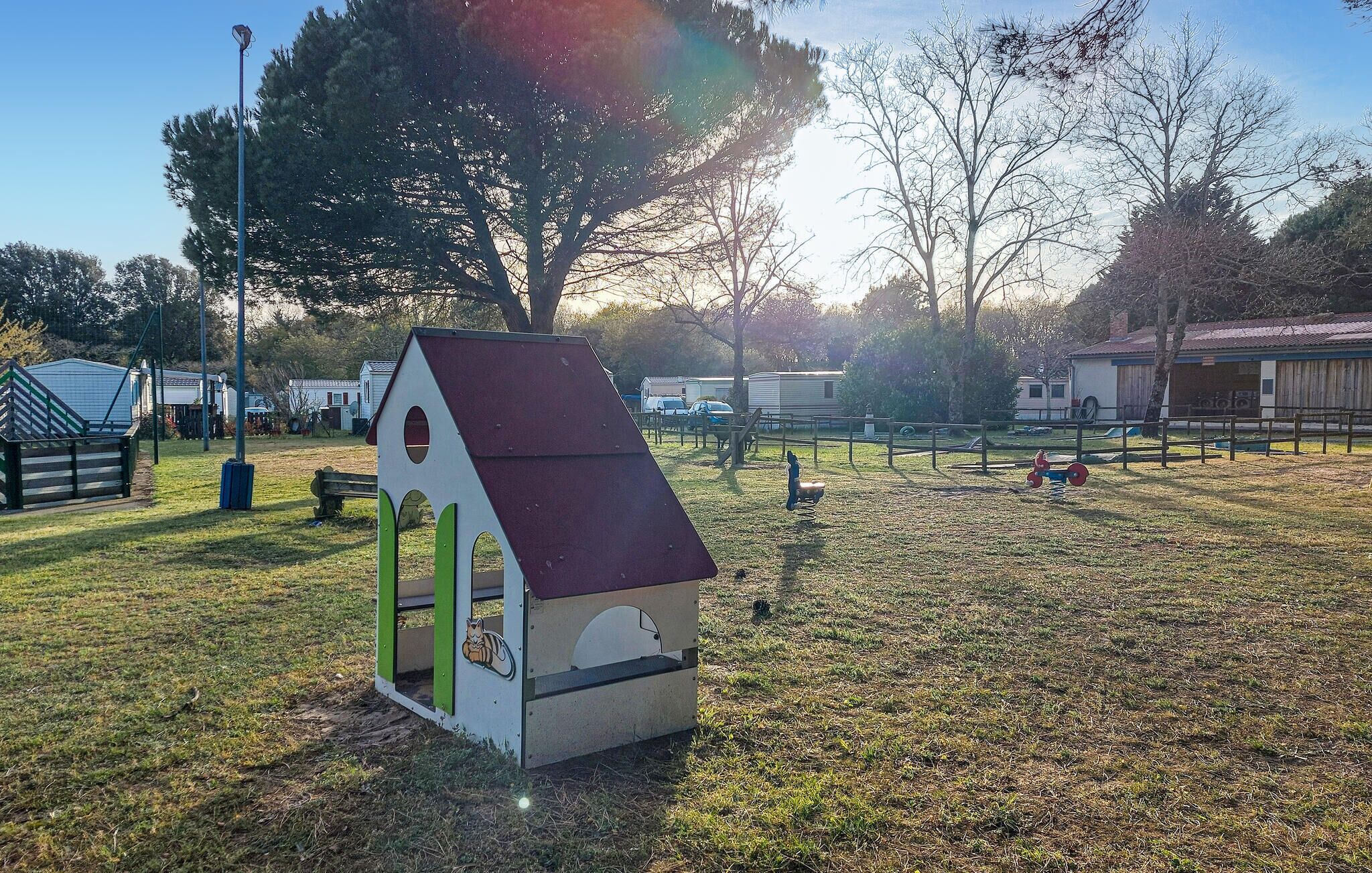 A small, triangular-roofed playhouse with a green door and a cat sticker, set in a grassy area with trees and a playground in the background.