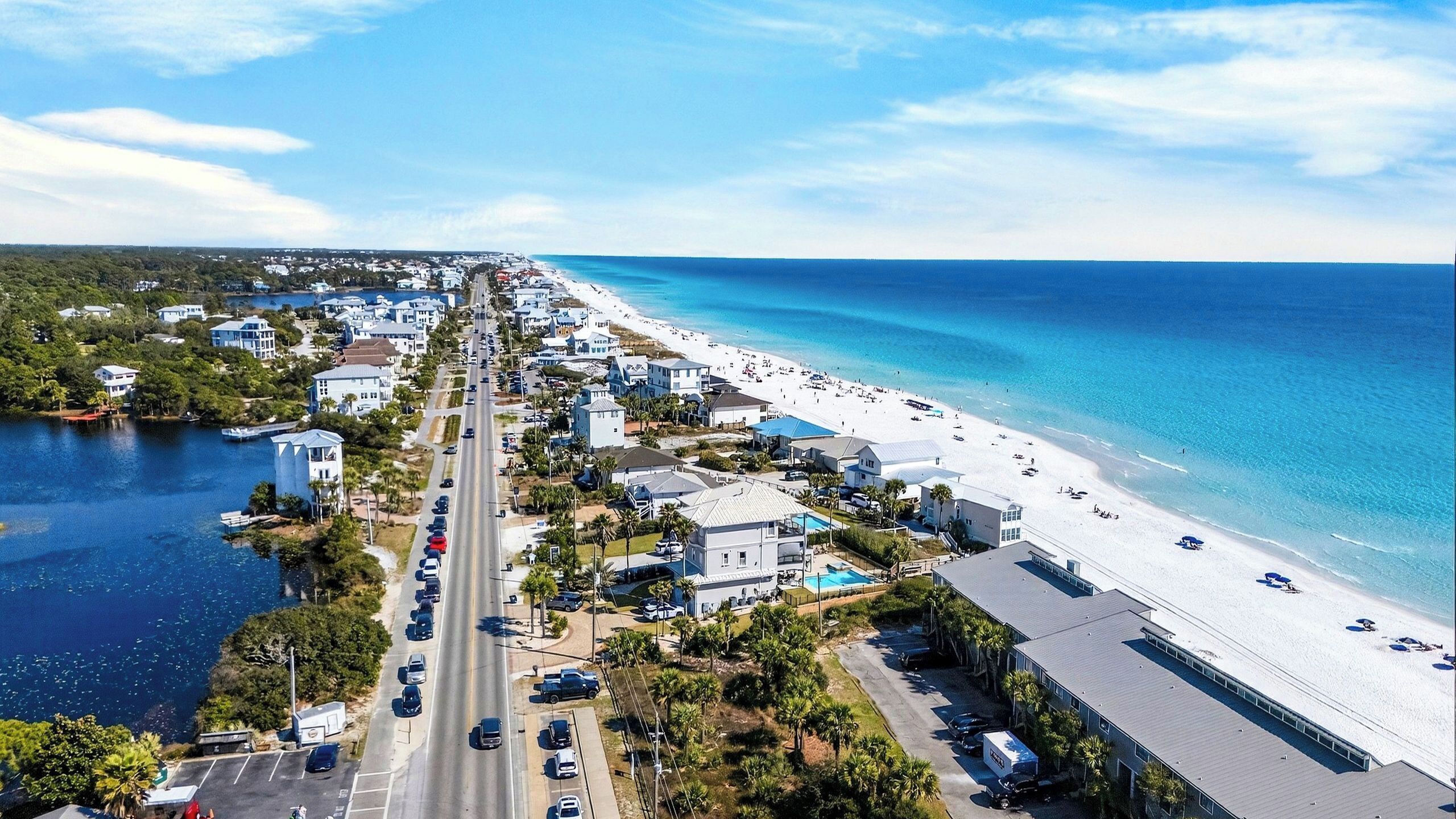 A coastal road with a beach on one side, lined with buildings and parked cars.