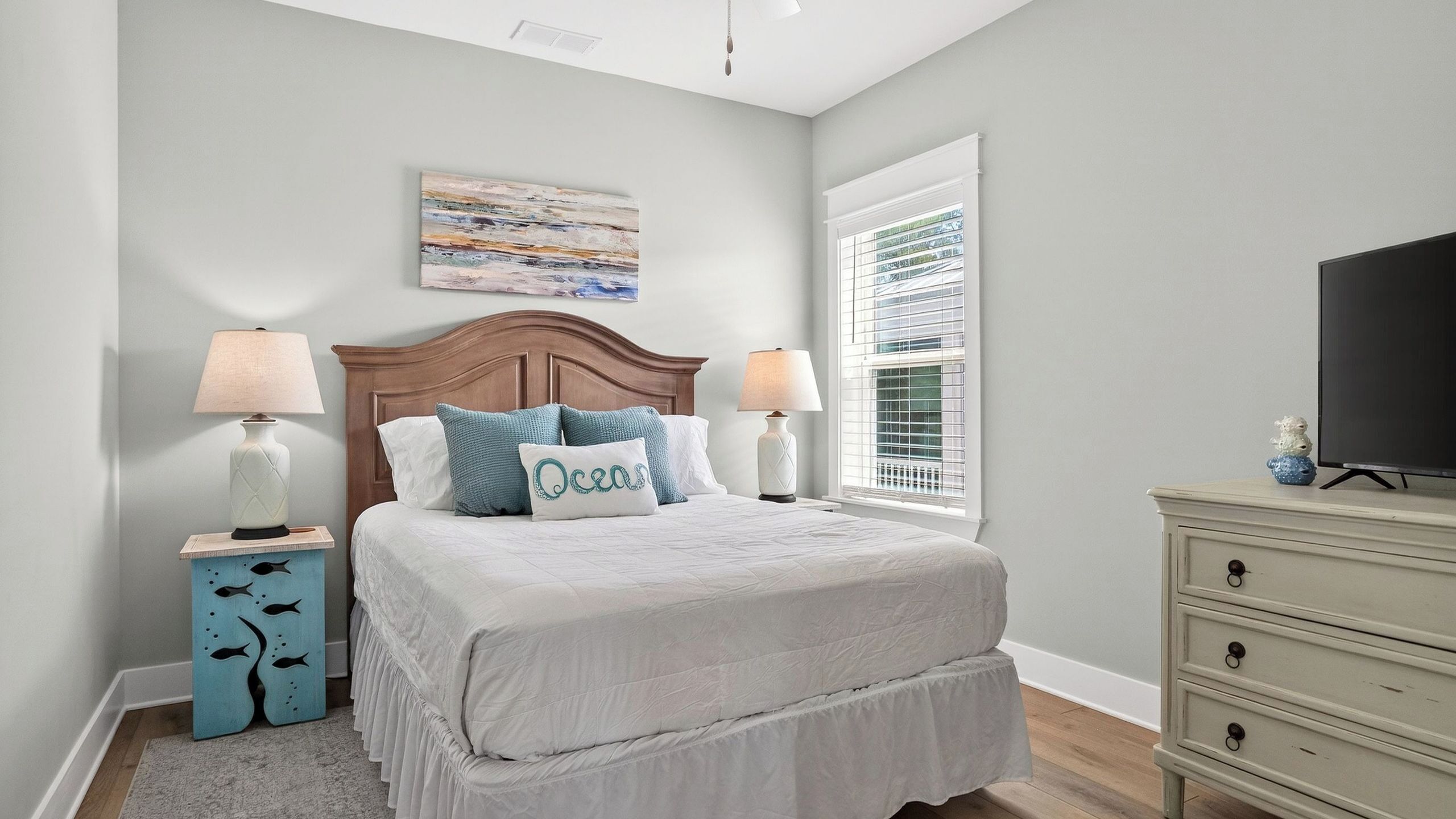 A bedroom with a bed, two bedside tables, a dresser, a television, a window with blinds, and a painting on the wall.