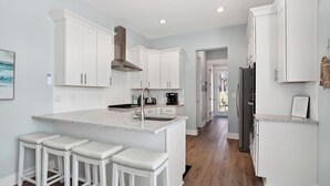 A modern kitchen with white cabinetry, a central island with bar stools, a stainless steel range hood, and a built-in oven. - Southern Dreams on 30A by Avantstay Pool + Outdoor Lounge + Golf Cart Access Minutes to the Beach (Santa Rosa Beach)