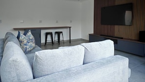 Ein modernes Wohnzimmer mit einer Couch, zwei Hocker, einem Flachbildfernseher, der an einer Wand aus Holzpaneelen angebracht ist, und einer Küchenzeile mit einer Arbeitsplatte und Hocker.