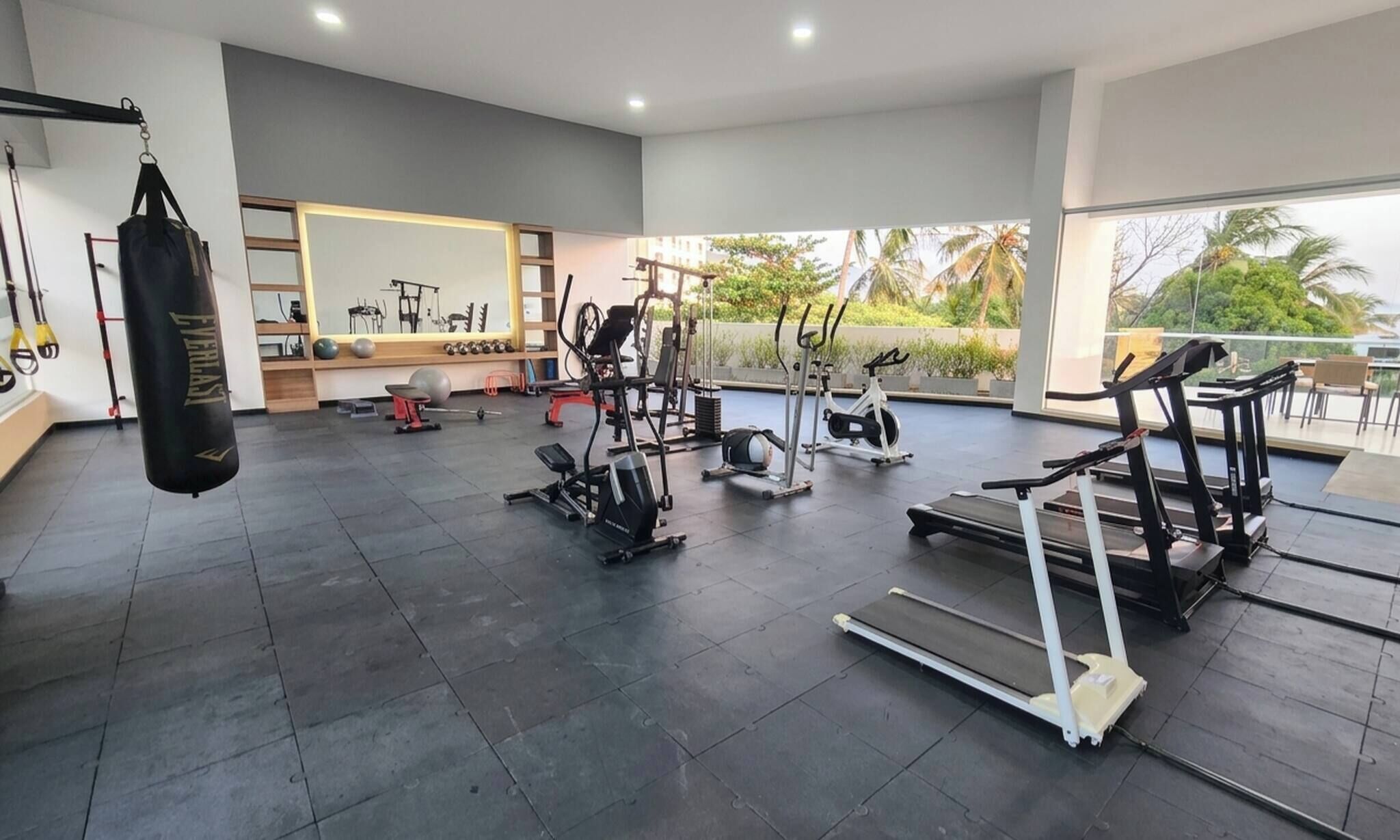 Un gimnasio amplio con diversos equipos de ejercicio, un saco de boxeo y grandes ventanales con vistas a palmeras.