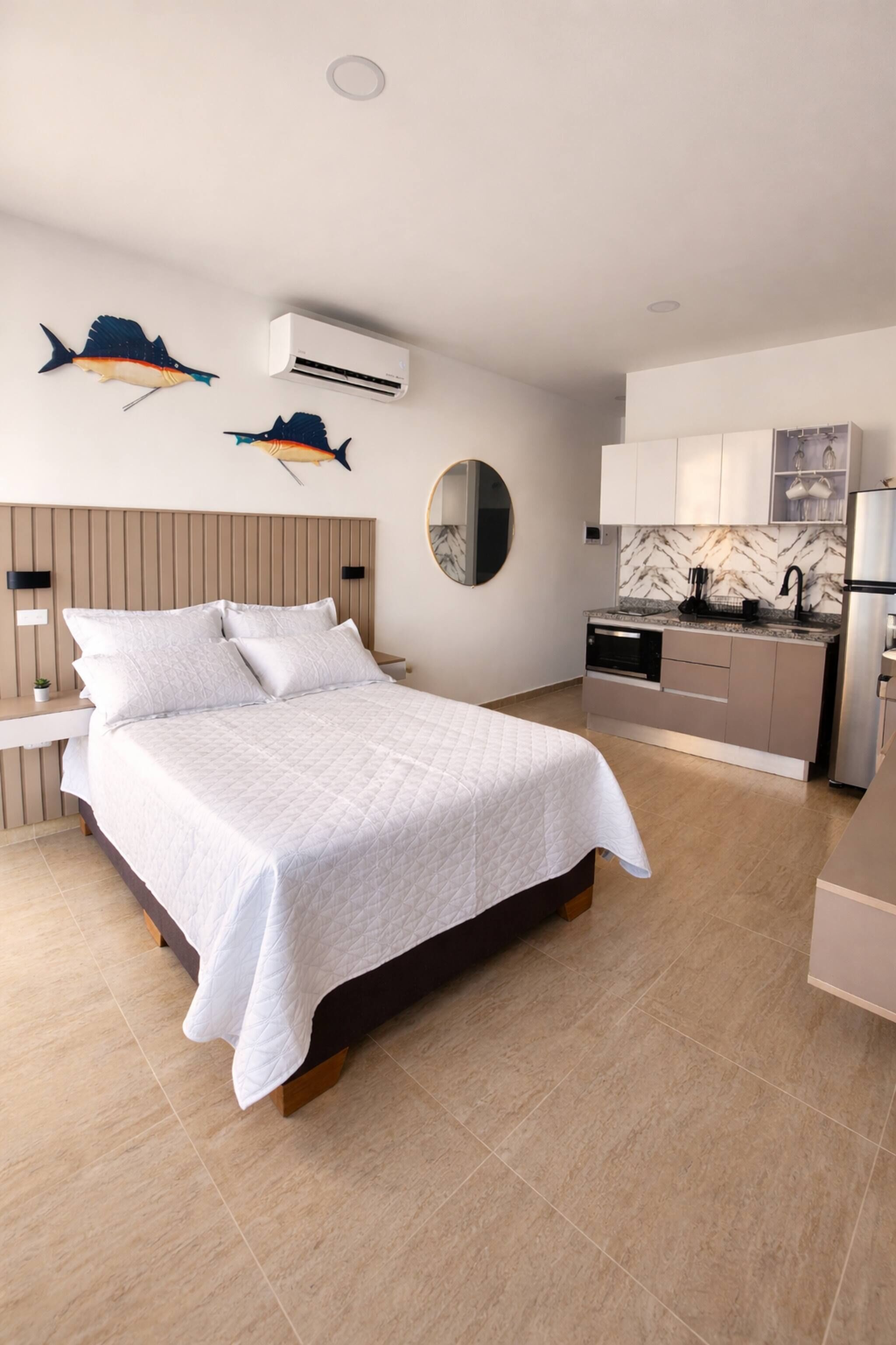 A modern bedroom with a large bed, a bedside table, a wall-mounted air conditioner, and a kitchen area with a marble backsplash.