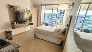 A modern hotel room with a large bed, a TV, a small desk, and a balcony with a view of the city. - Ocean View and Direct Beach in Pozos Colorados (Santa Marta)