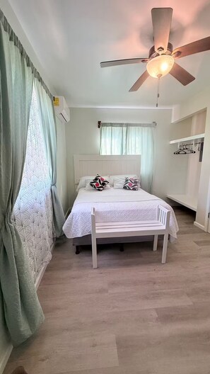 Ein Schlafzimmer mit einem Bett, einem Deckenventilator, einem Fenster mit Vorhängen und einem an der Wand befestigten Regal.