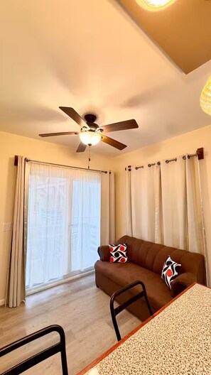 Ein Zimmer mit Deckenventilator, einem braunen Sofa mit gemusterten Kissen, einem Esstisch mit einem Stuhl und einem großen Fenster mit Vorhängen.