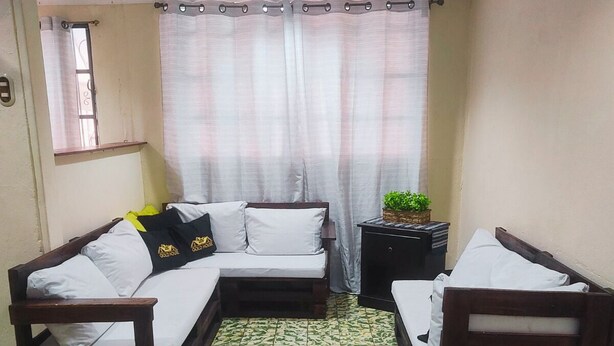 Ein Zimmer mit einer Couch, zwei Stühlen, einem kleinen Tisch mit Pflanze und einem Fenster mit Vorhängen.