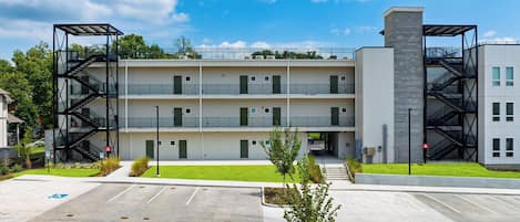 Un edificio de varios pisos con escaleras de incendios exteriores, un estacionamiento y una pequeña zona verde con árboles.