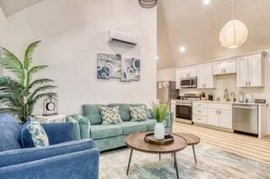 A living room with a sofa, a coffee table, and a plant. There is a kitchen with white cabinets and appliances. - 2 Private Cabins • 2 Hot Tubs • Sleeps 8 (Sevierville)