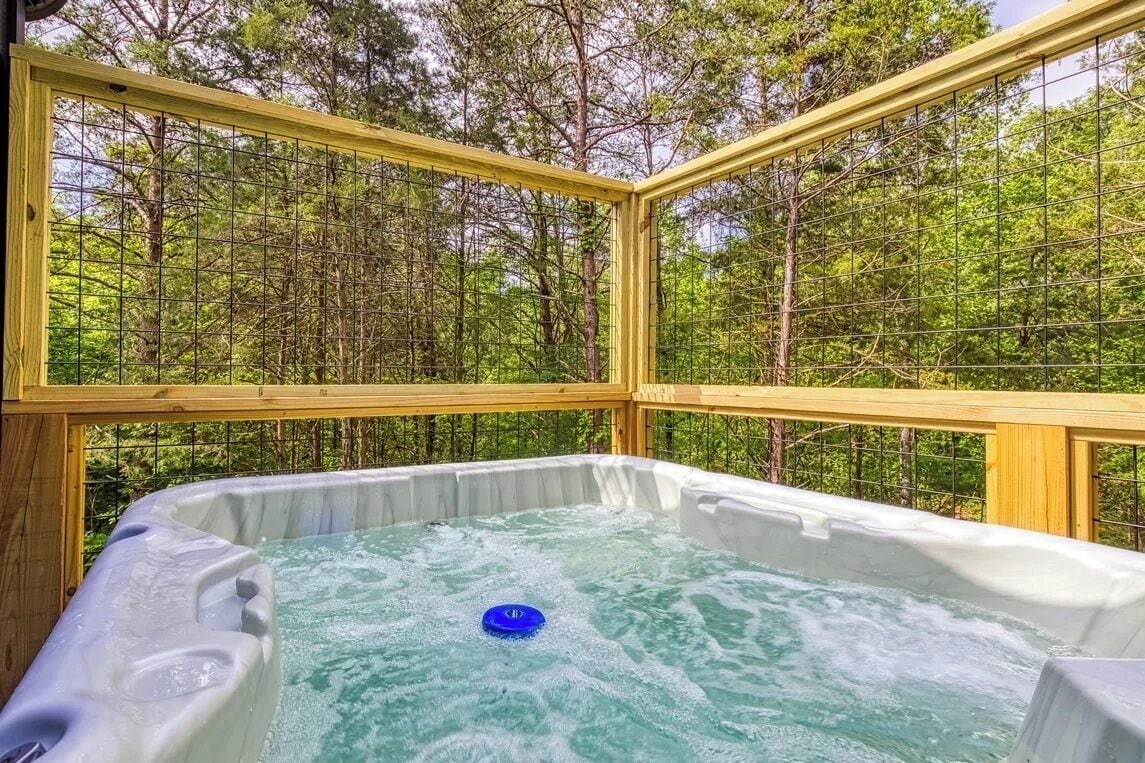 Een jacuzzi met een blauw drijvend object, omgeven door een houten terras en een hek van gaas, met een bos op de achtergrond.